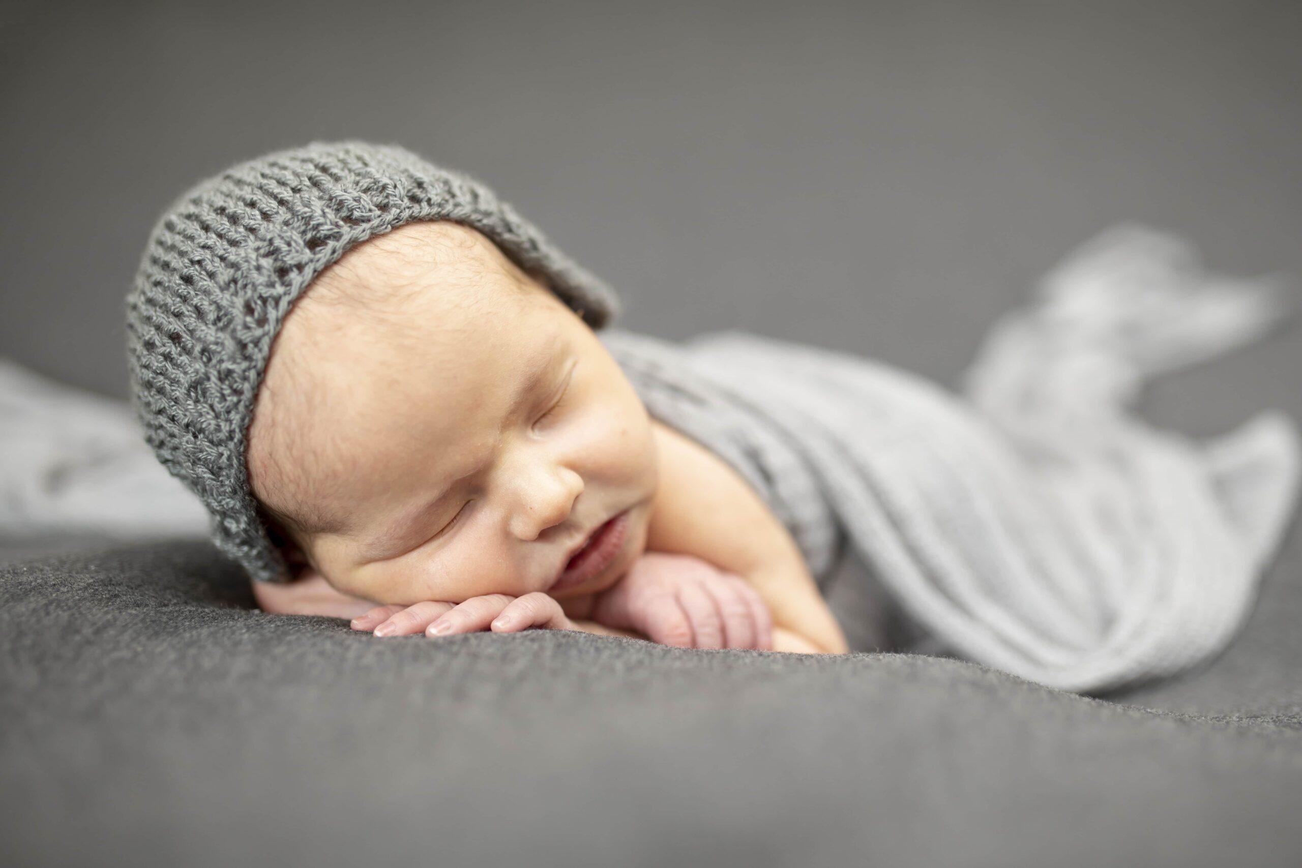 newborn sleeping on a gray backdrop with a gray bonnet on his head