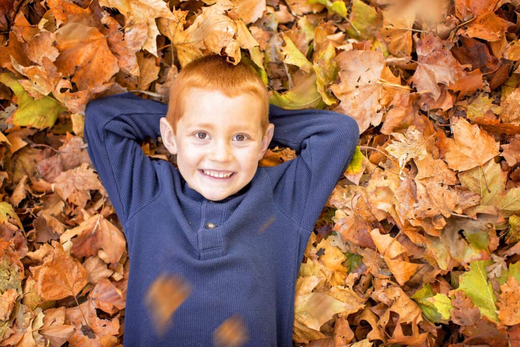 A young boy laying in a pile of fall leaves looking at the camera