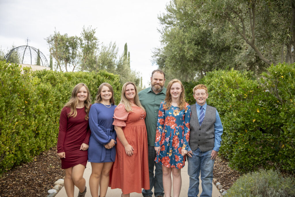 Family of six stand together with green hedges behind them