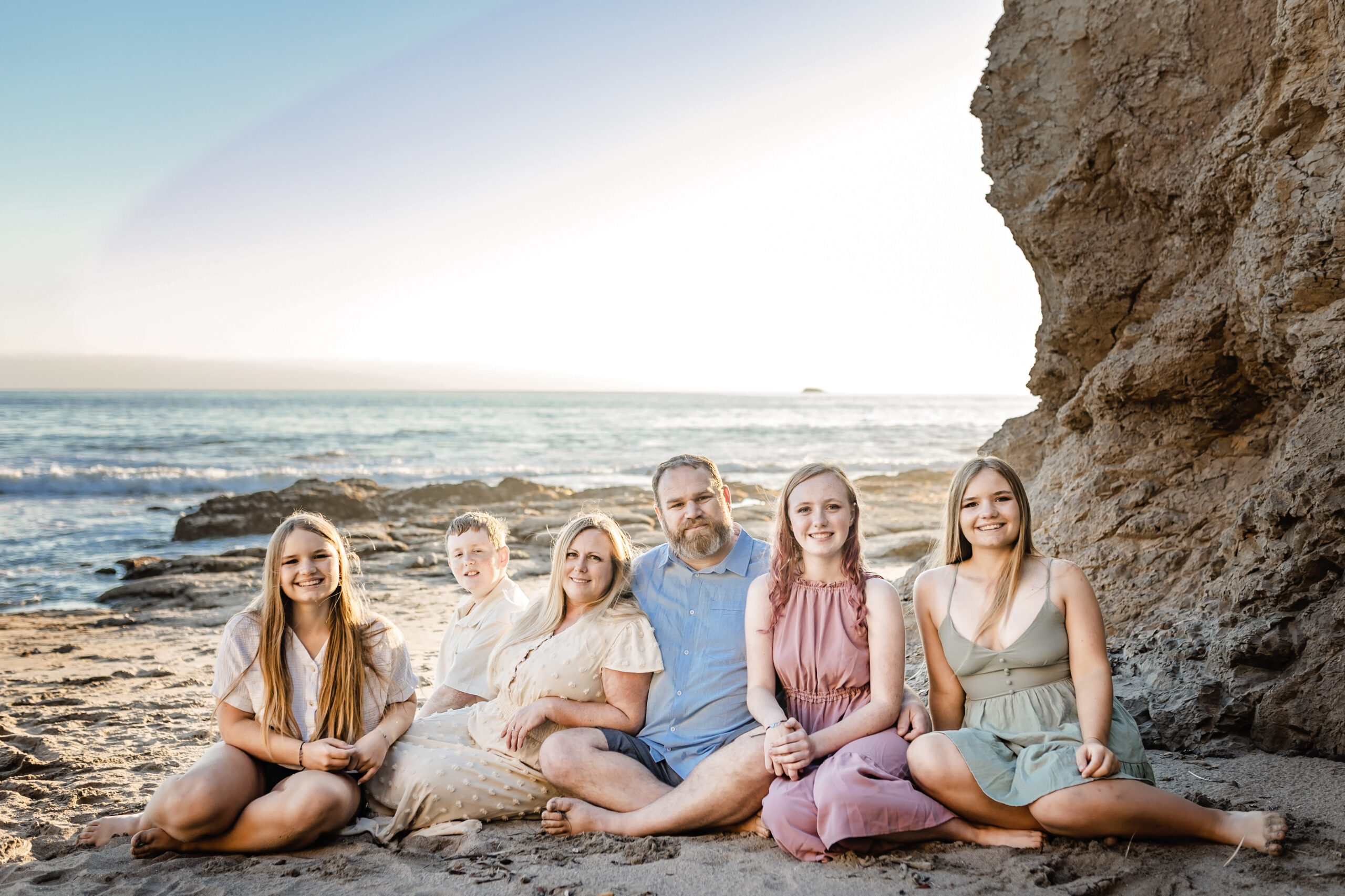 Family of six sitting in the sand at the beach with the ocean behind them.