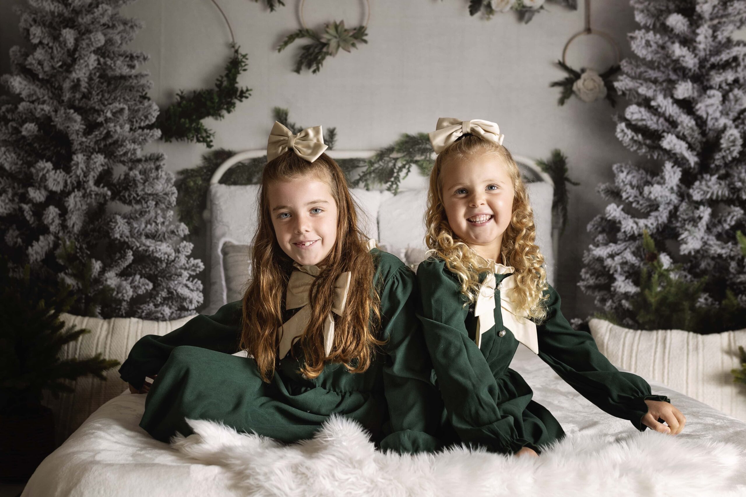 Two girls sitting together in green dresses on a bed with a Christmas themed background