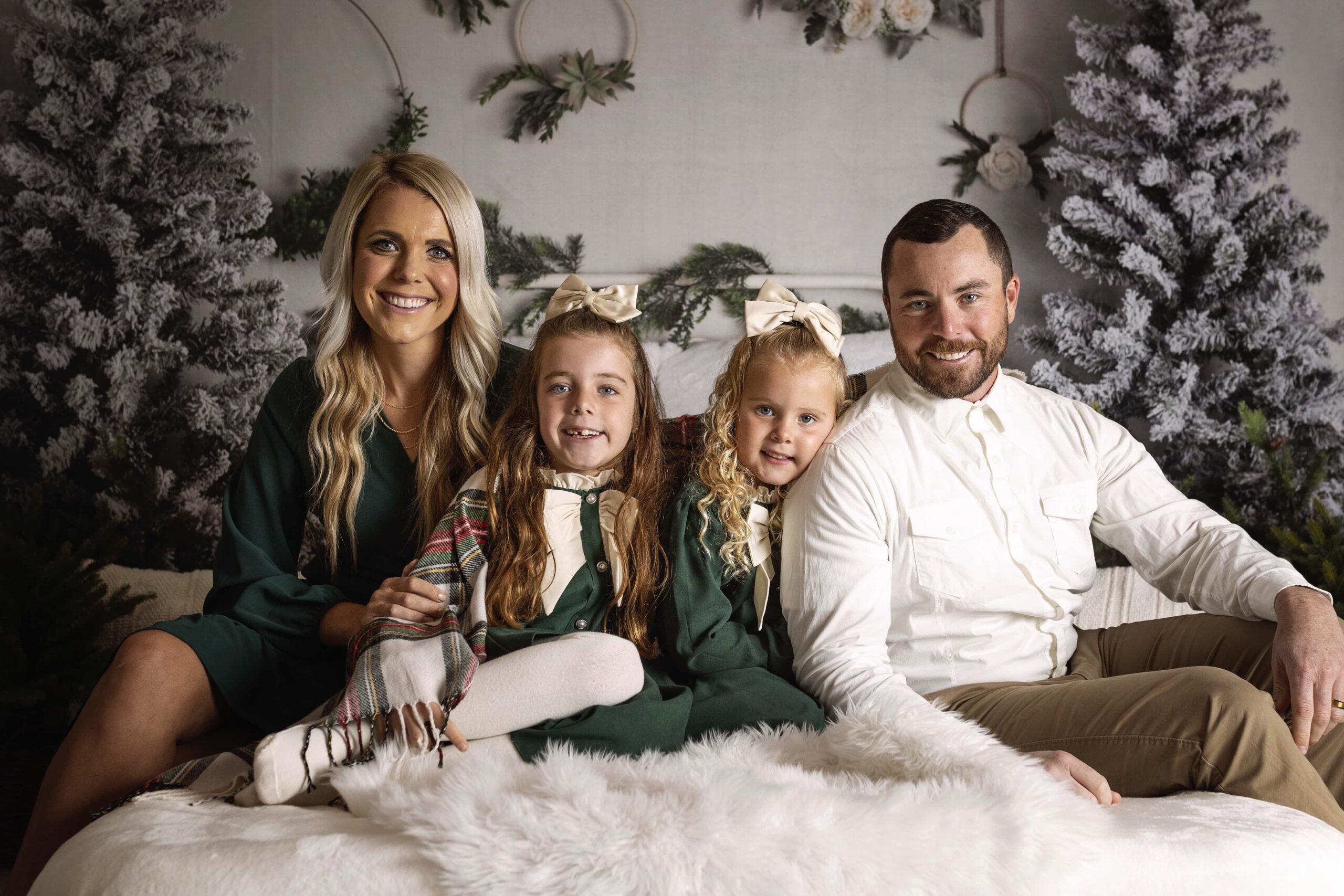 family of four with two young girls sitting on a Christmas themed bed during a mini session