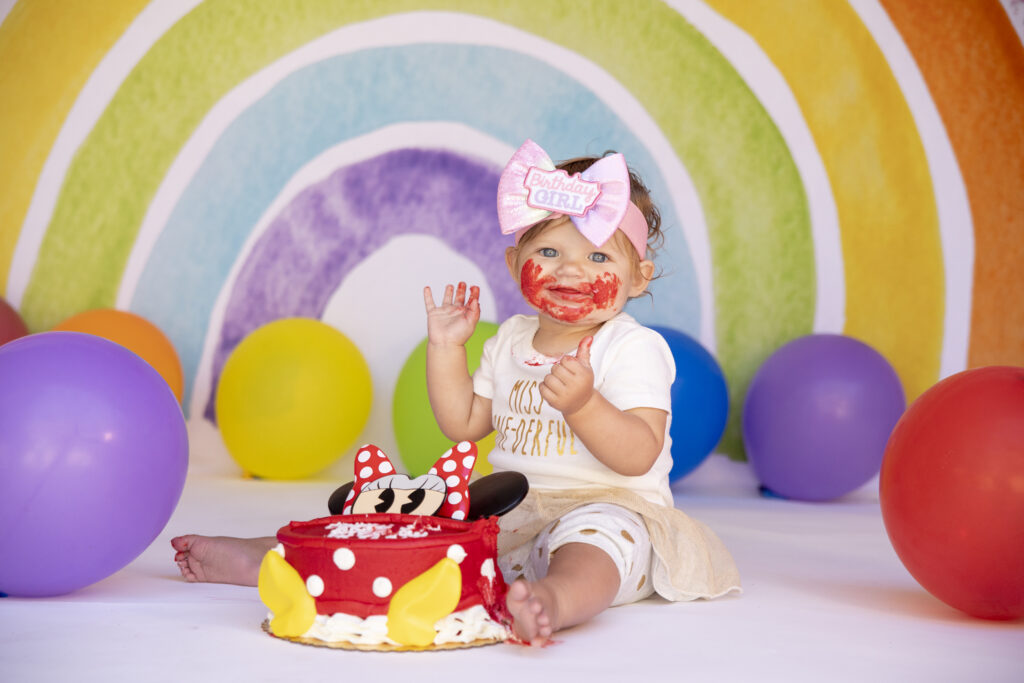 One year old girl tasting her birthday cake in front of a rainbow backdrop