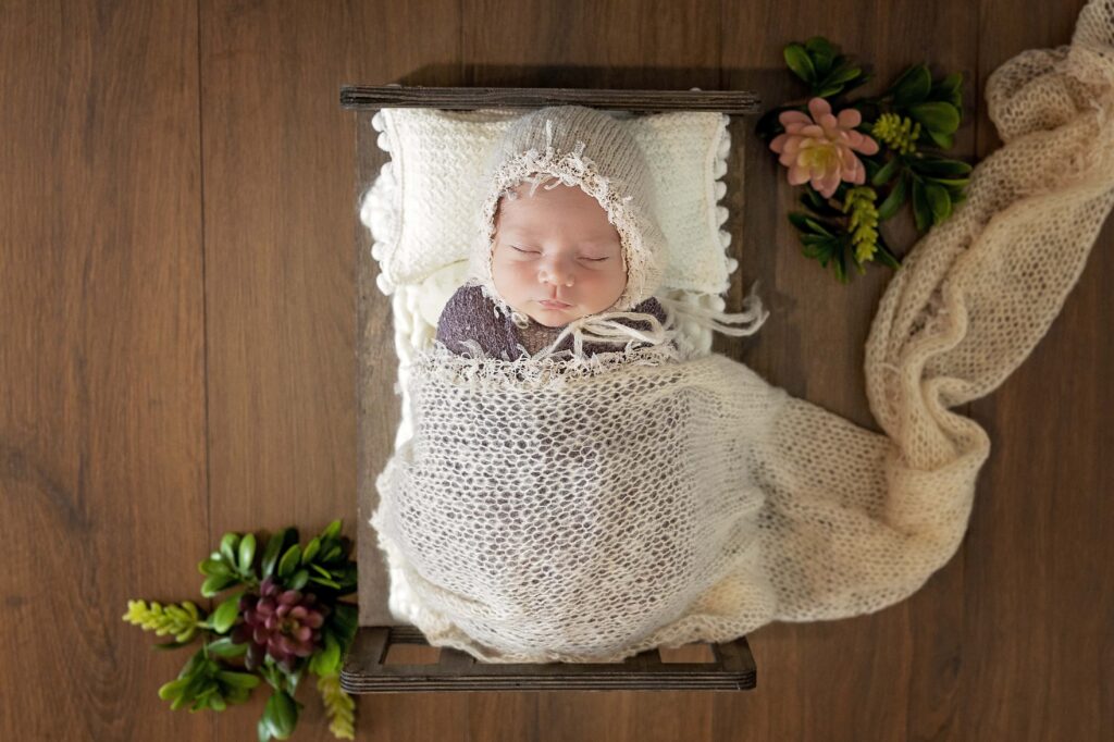 newborn girl on a doll bed wearing a bonnet with greenery and succulents around her