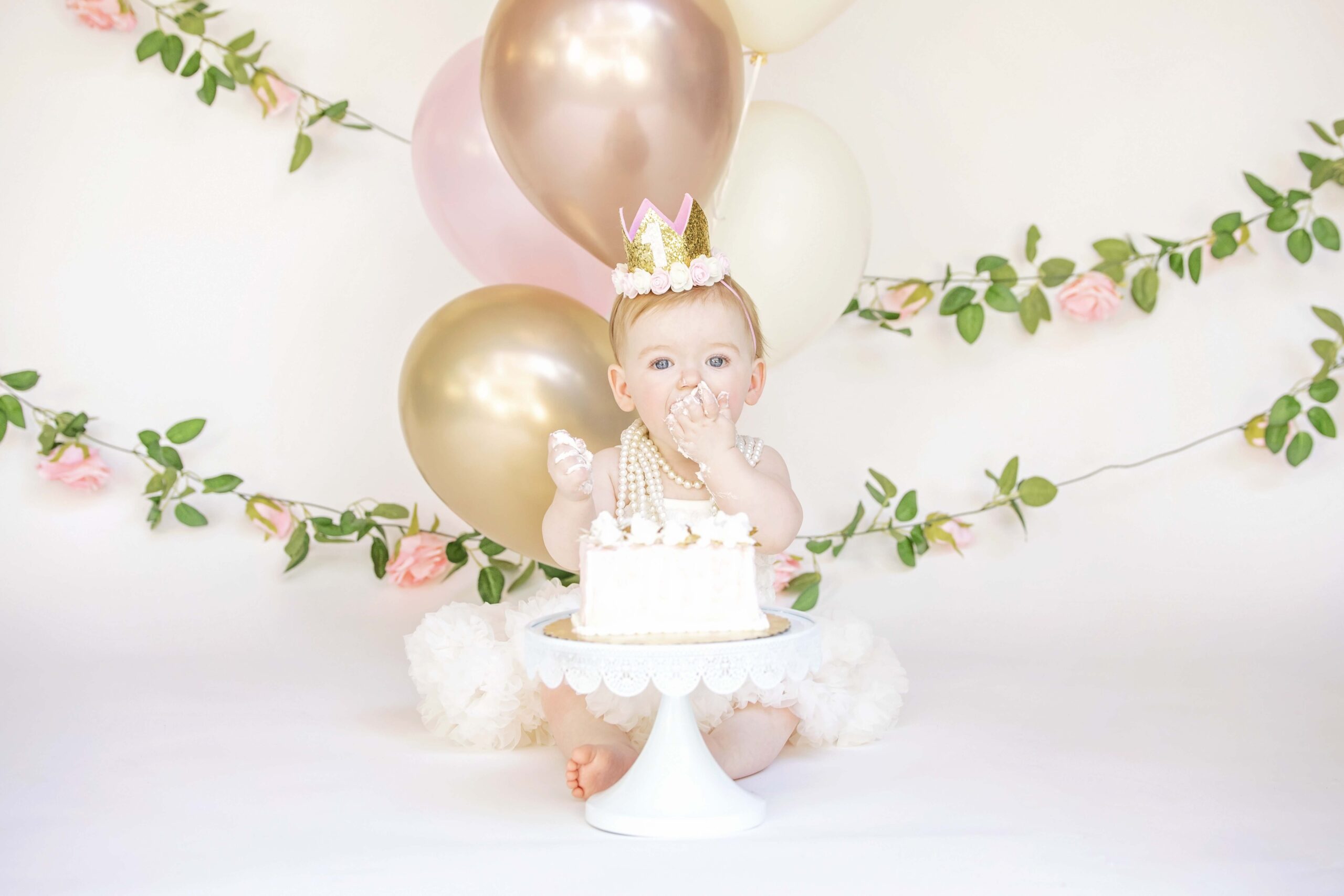 One year old girl tasting her birthday cake in front of gold balloons and a floral garland