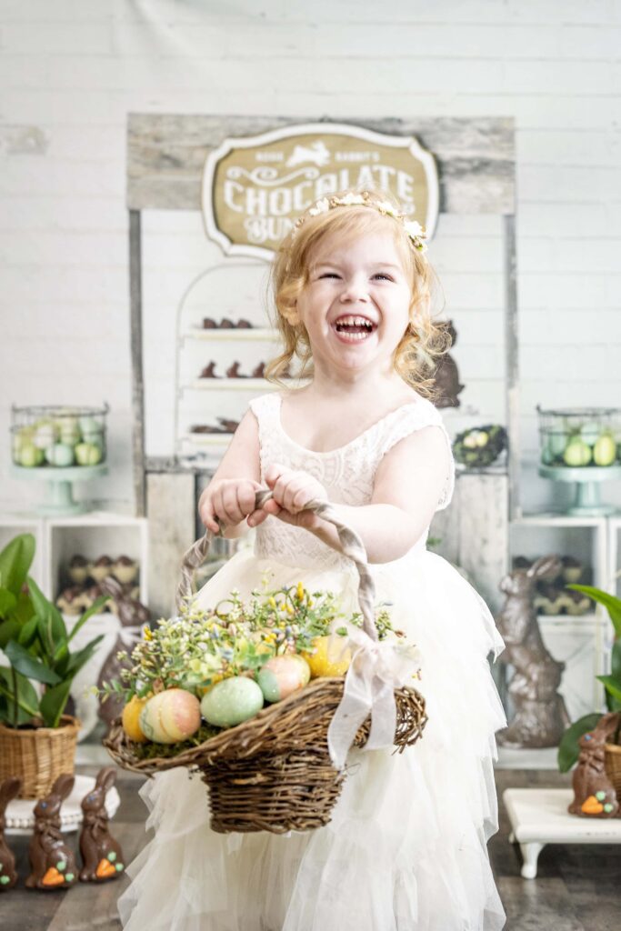 young girl in a white dress holding a basket of easter eggs with a chocolate easter bunny themed background