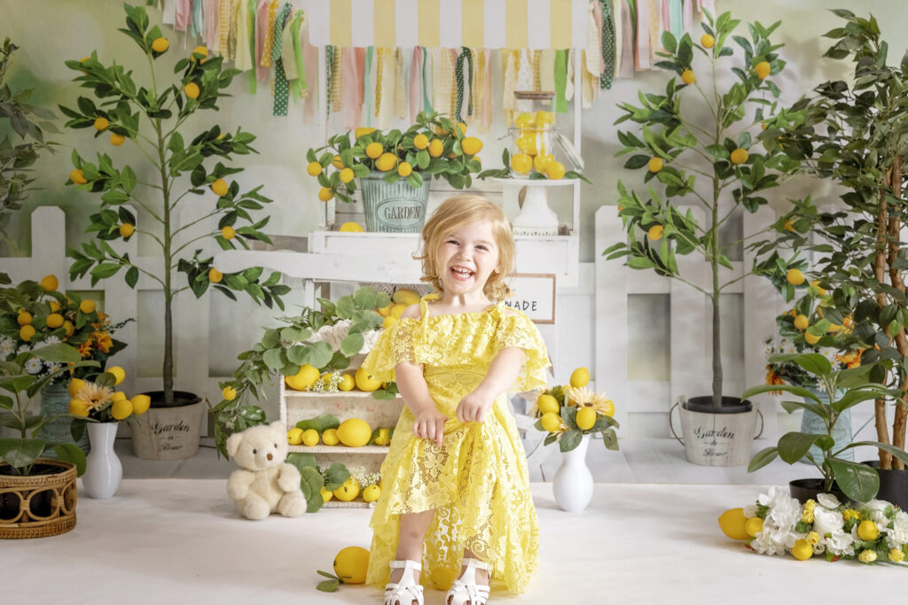 3 year old girl in a yellow lace dress sitting on a chair with a lemonade stand background