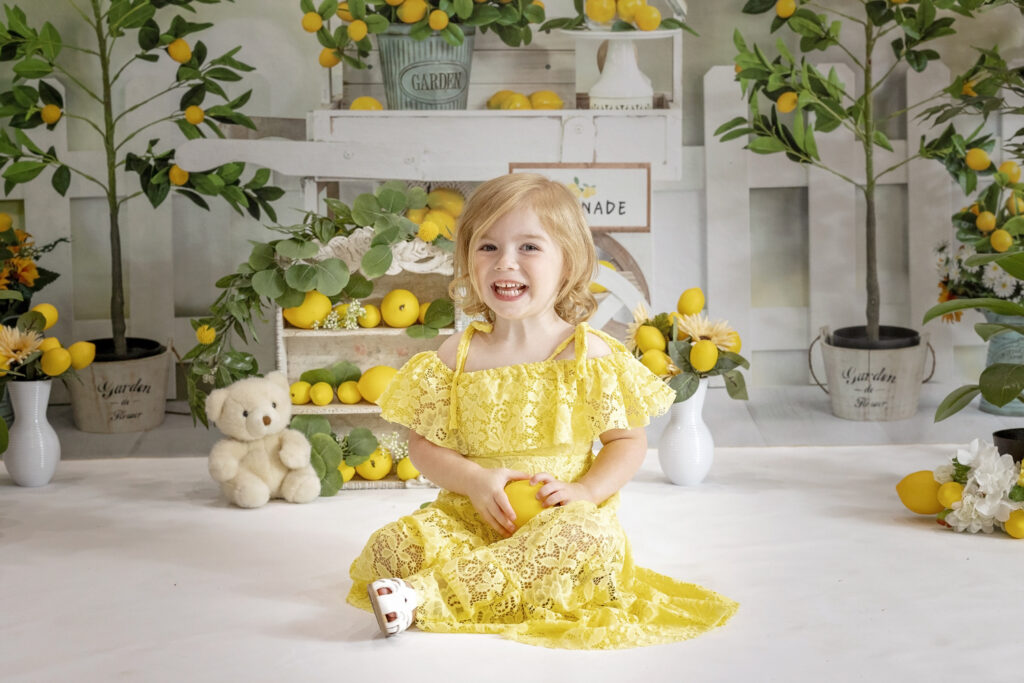 young girl in a yellow lace dress sitting on a white floor holding a lemon with a lemonade stand background