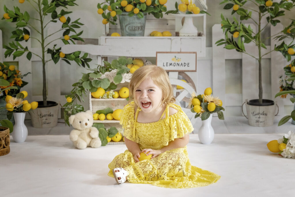 young girl laughing at the camera in a yellow lace dress sitting on a white floor holding a lemon with a lemonade stand background