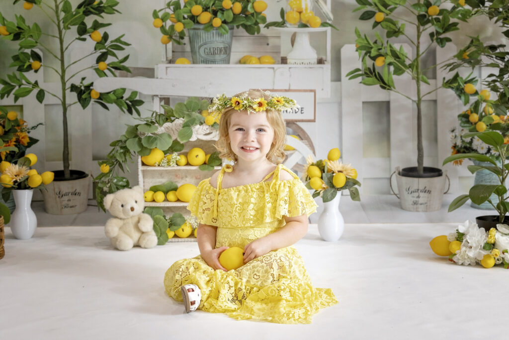 young girl in a yellow lace dress and a flower crown sitting on a white floor with a lemonade stand background