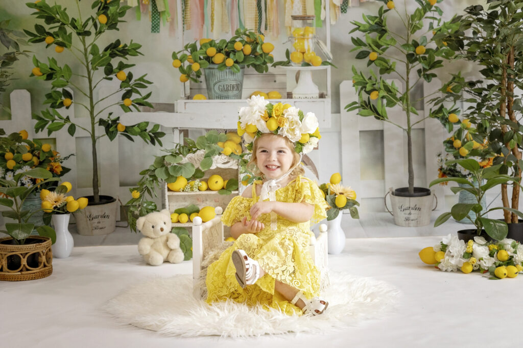 young girl in a yellow lace dress and a flower crown sitting on a white doll bed with a lemonade stand background