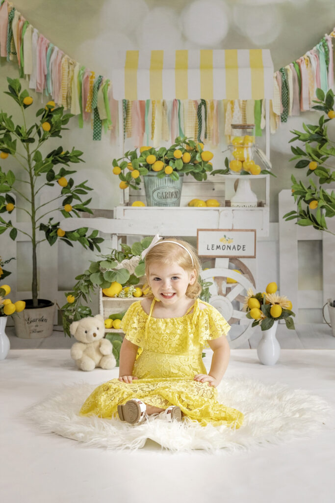 young girl in a yellow lace dress sitting on a white rug with a lemonade stand background