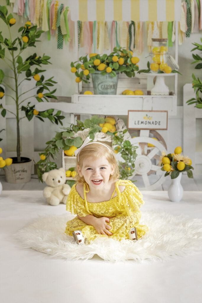 young girl in a yellow lace dress sitting on a white rug with a lemonade stand background