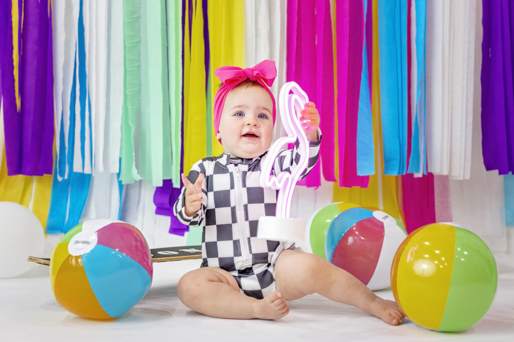 one year old girl in a checkered outfit holding a light up flamingo with colorful paper streamers in the background