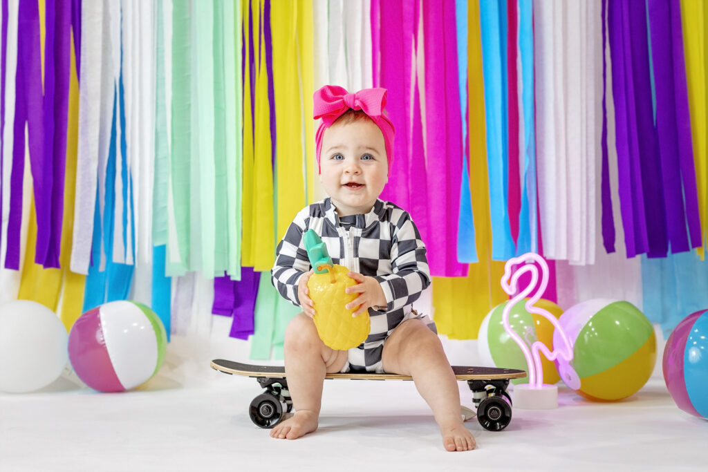 one year old girl in a black and white checkered outfit sitting on a skateboard, holding a pineapple cup with beach balls and a colorful streamer background