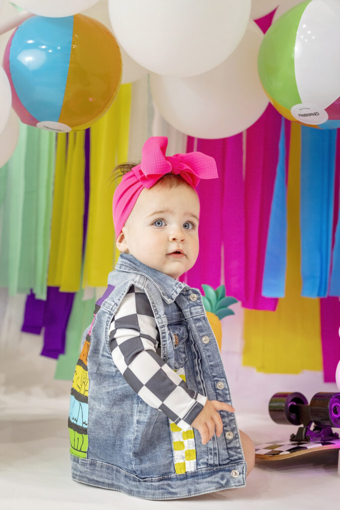 one year old girl in a jean vest looking back at the camera with a colorful streamer background with balloons and beach balls