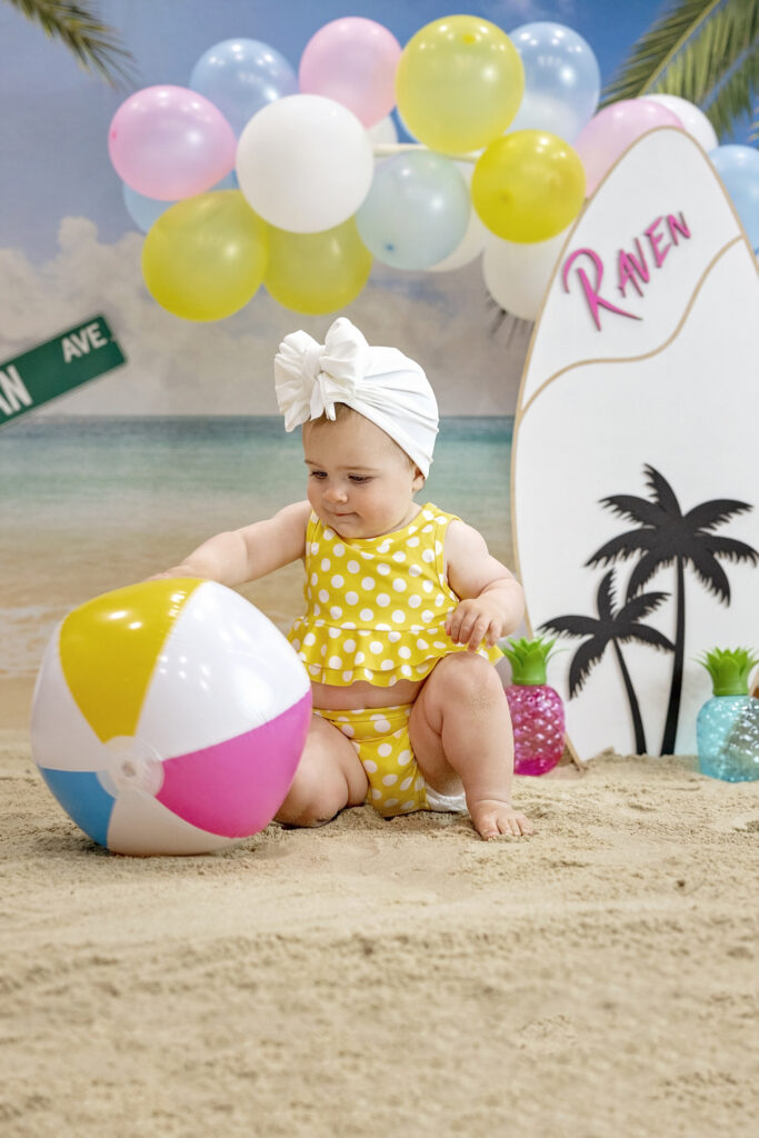 one year old girl in a yellow bathing suit sitting in the sand with a beach background and a wood surfboard and a beach ball