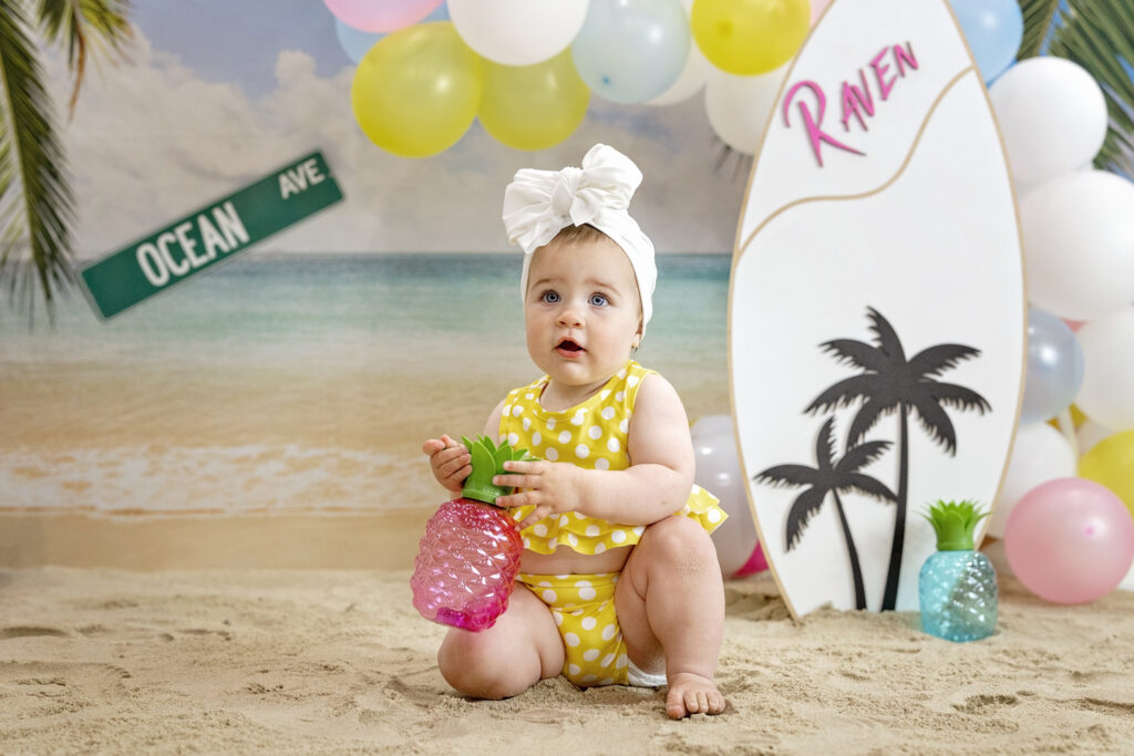 one year old girl in a yellow bathing suit sitting in the sand with a beach background and a wood surfboard holding a pink pineapple cup