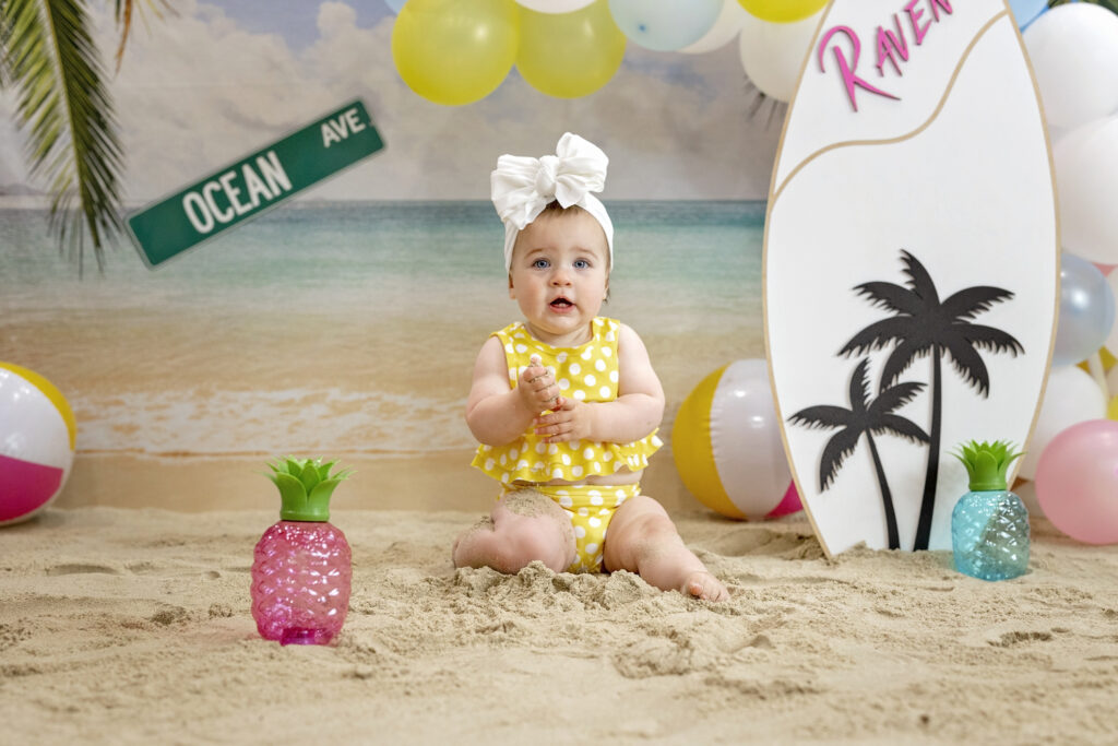 one year old girl in a yellow bathing suit sitting in the sand with a beach background and a wood surfboard