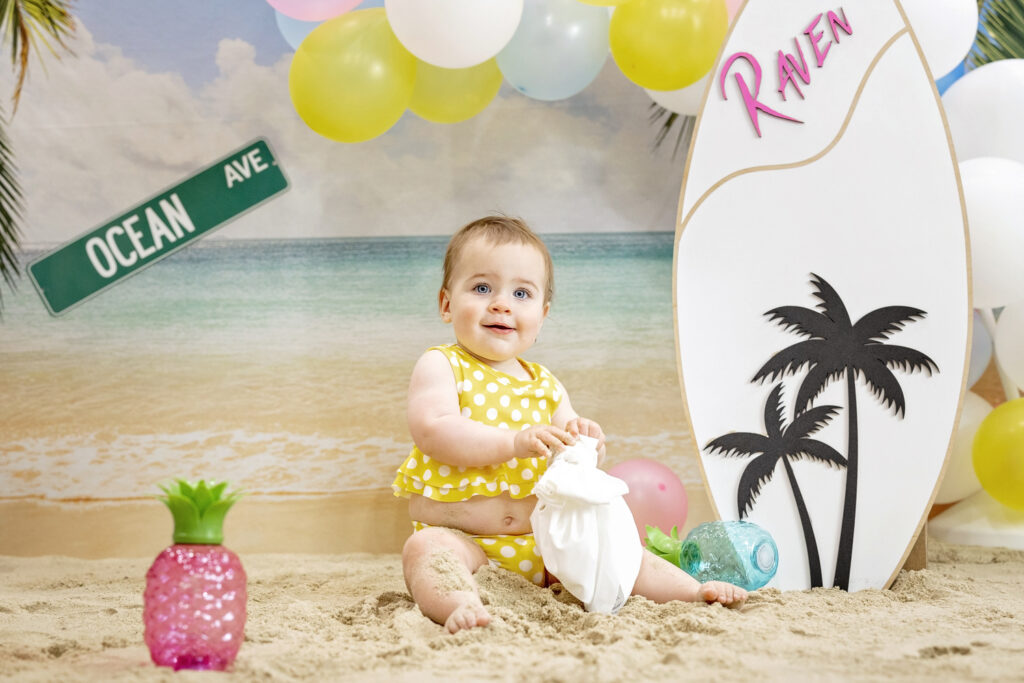 one year old girl in a yellow bathing suit sitting in the sand with a beach background and a wood surfboard