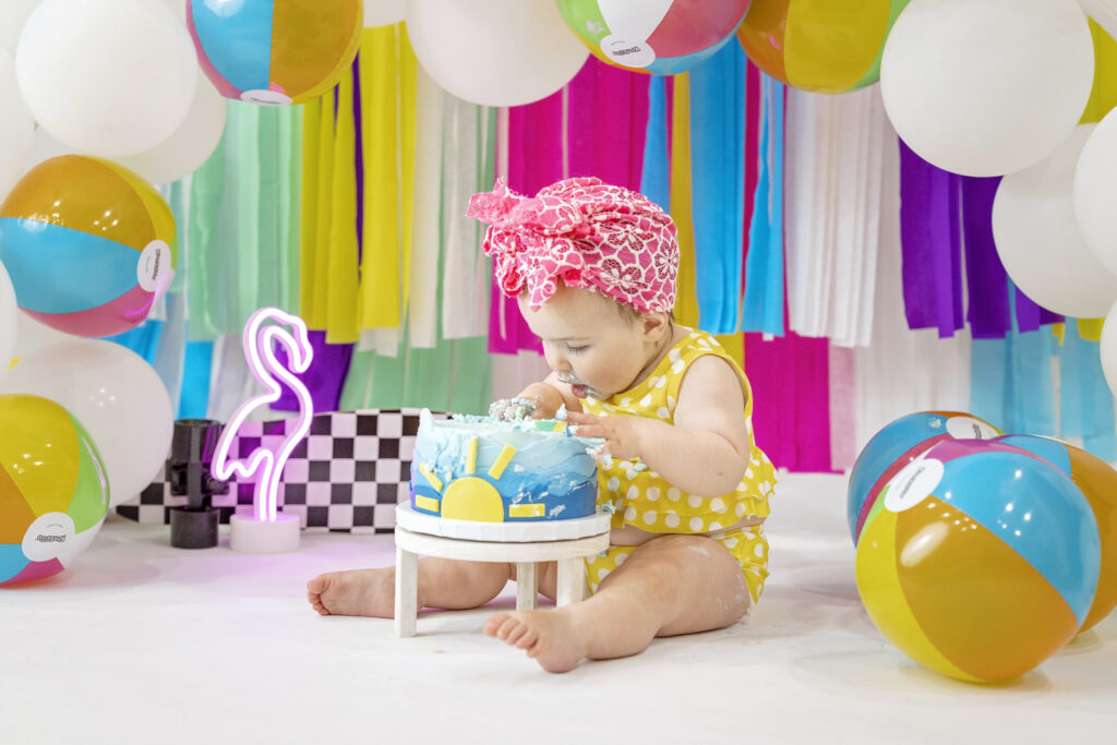 one year old girl in a yellow bathing suit with a colorful streamer background tasting the frosting from her first birthday cake in shades of blue with a yellow sun