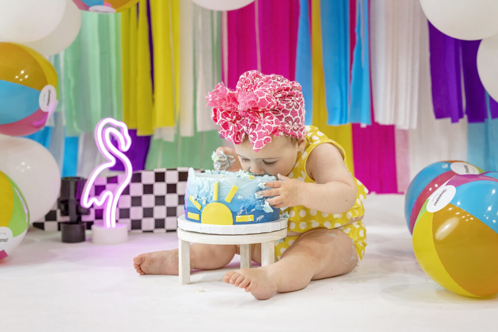 one year old girl in a yellow bathing suit with a colorful streamer background putting her face in the birthday cake in shades of blue with a yellow sun