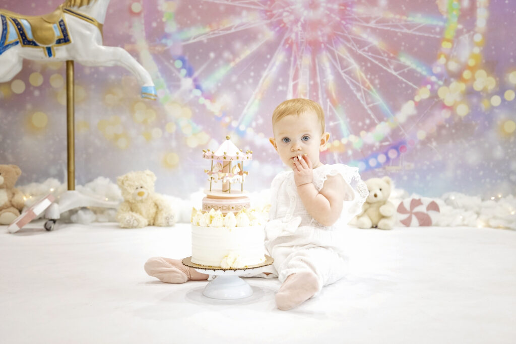 one year old girl in a white romper eating a white cake in front of a pink carnival background