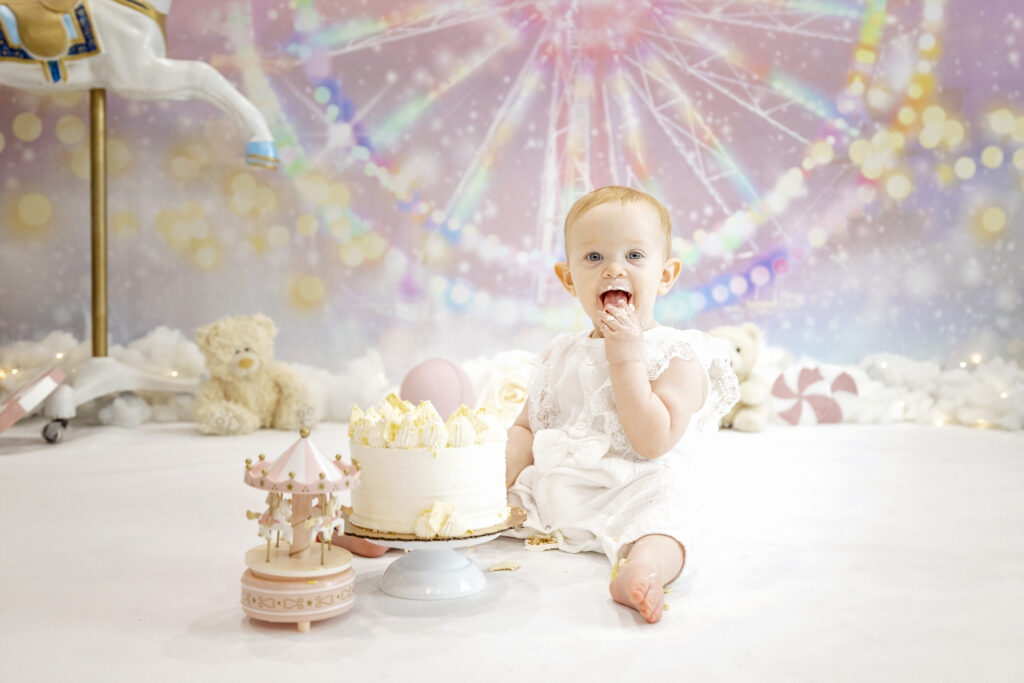 one year old girl in a white romper eating a white cake in front of a pink carnival background