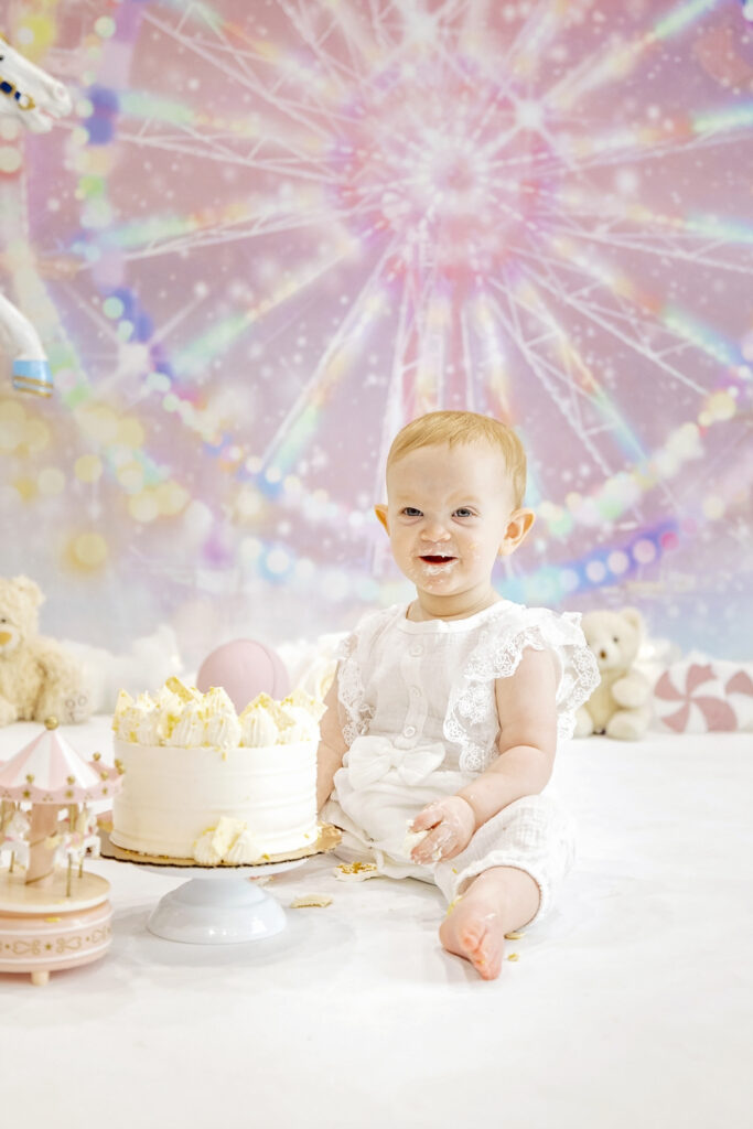 one year old girl in a white romper eating a white cake in front of a pink carnival background