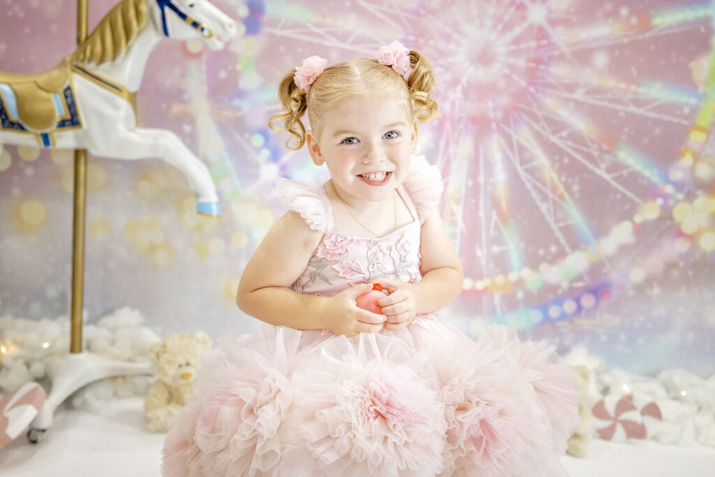 4 year old girl in a pink fluffy dress sitting in front of a pink carnival background