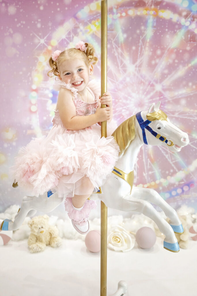 4 year old girl in a pink fluffy dress sitting on a carousel horse in front of a pink carnival background
