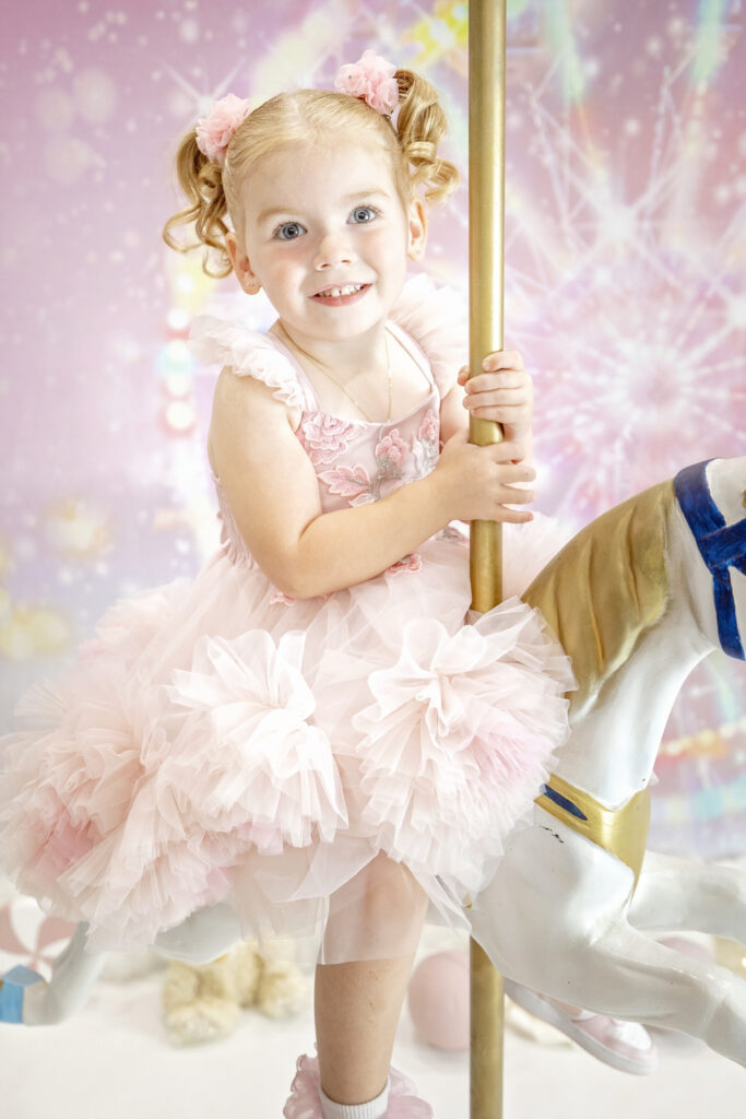 4 year old girl in a pink fluffy dress sitting on a carousel horse in front of a pink carnival background