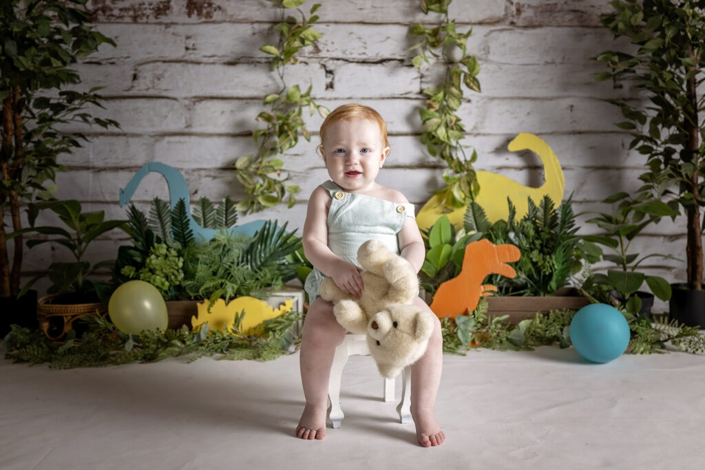 smiling one year old boy sitting in a white chair with a dinosaur theme behind him