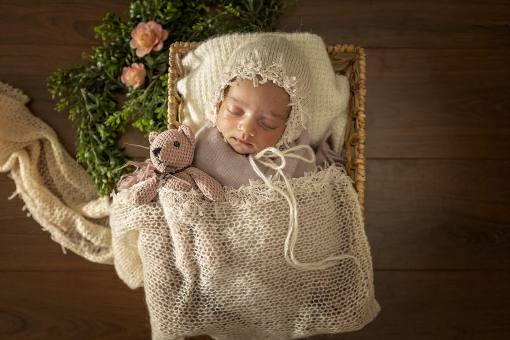 Newborn girl in a basket with a white bonnet and a pink teddy bear