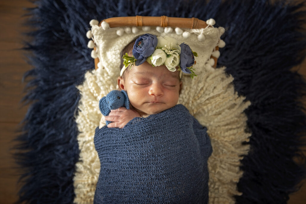 Newborn girl on a boho bed with a blue wrap and blue flower headband and a blue tiny elephant