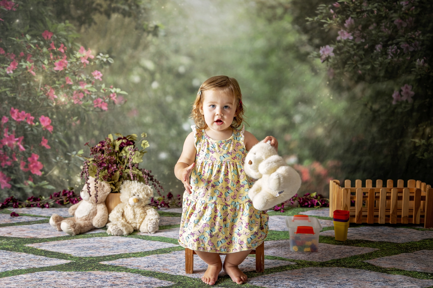one year old girl sitting on a small chair in a butterfly dress holding a teddy bear