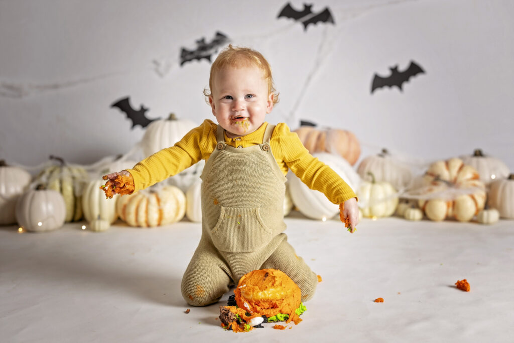 one year old boy in a yellow romper putting his hands on an orange pumpkin cake