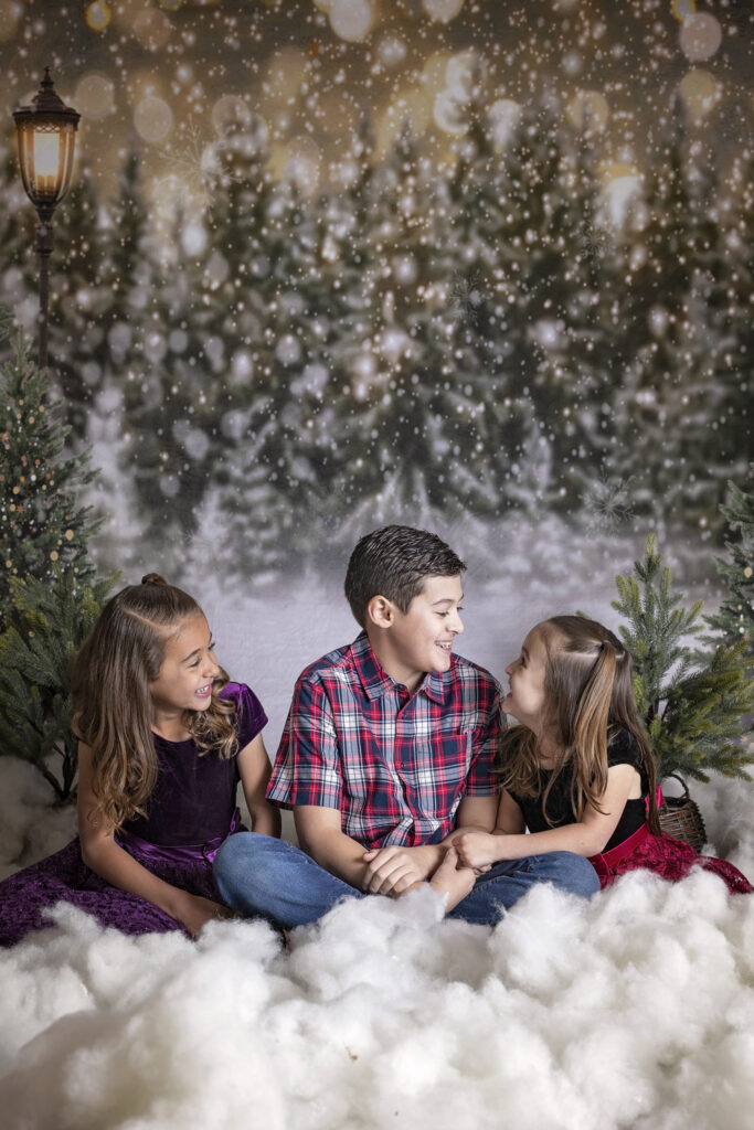 three siblings looking at each other while sitting on the floor in a Christmas tree farm studio set