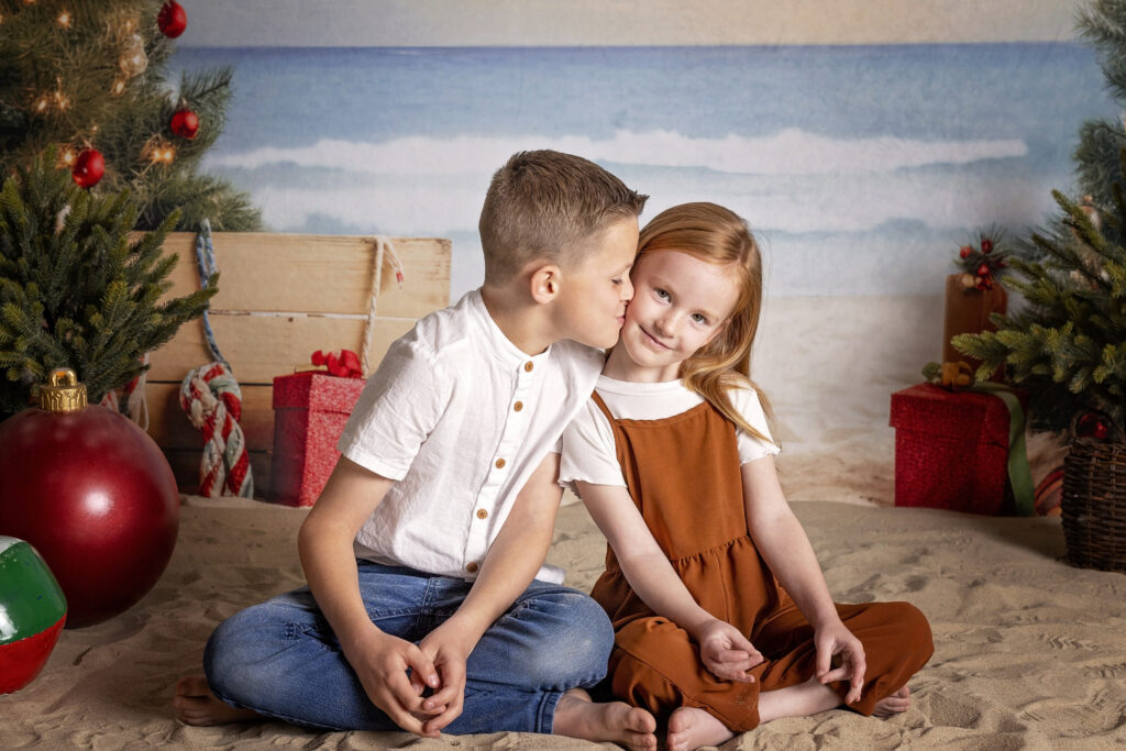 young boy kissing his sister on the cheek while they sit together in the sand