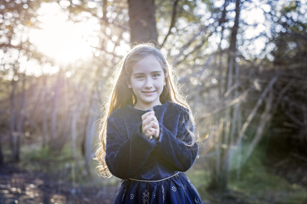 young girl in a blue dress clasping her hands together with the sun behind her