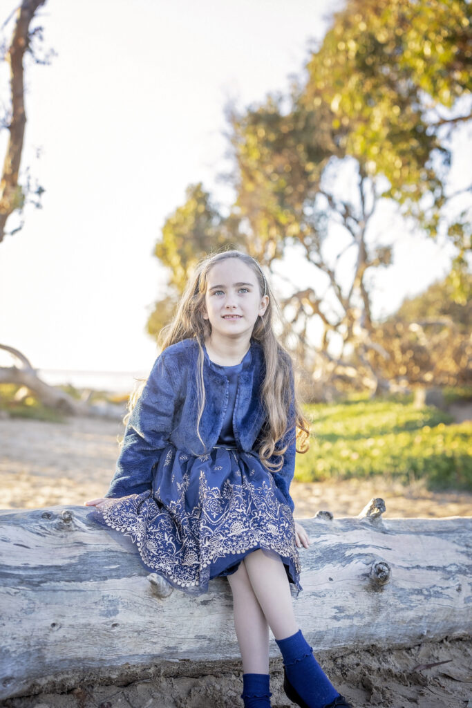 young girl in a blue dress sitting on a log at the beach