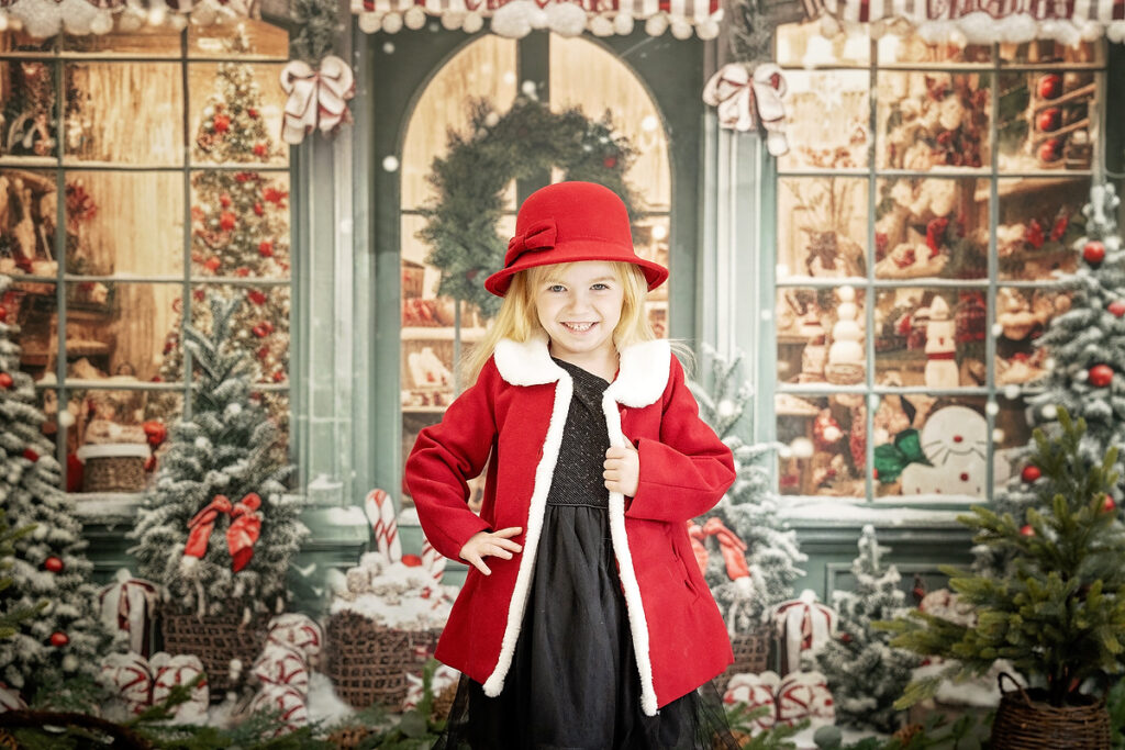 young girl in a black dress with a red coat in front of a Christmas themed storefront.