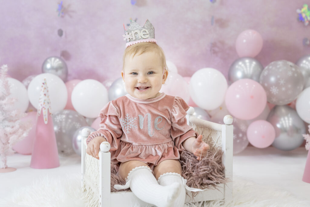 one year old girl in a pink dress smiling at the camera while sitting on a white doll bed with pink and silver balloons in the background