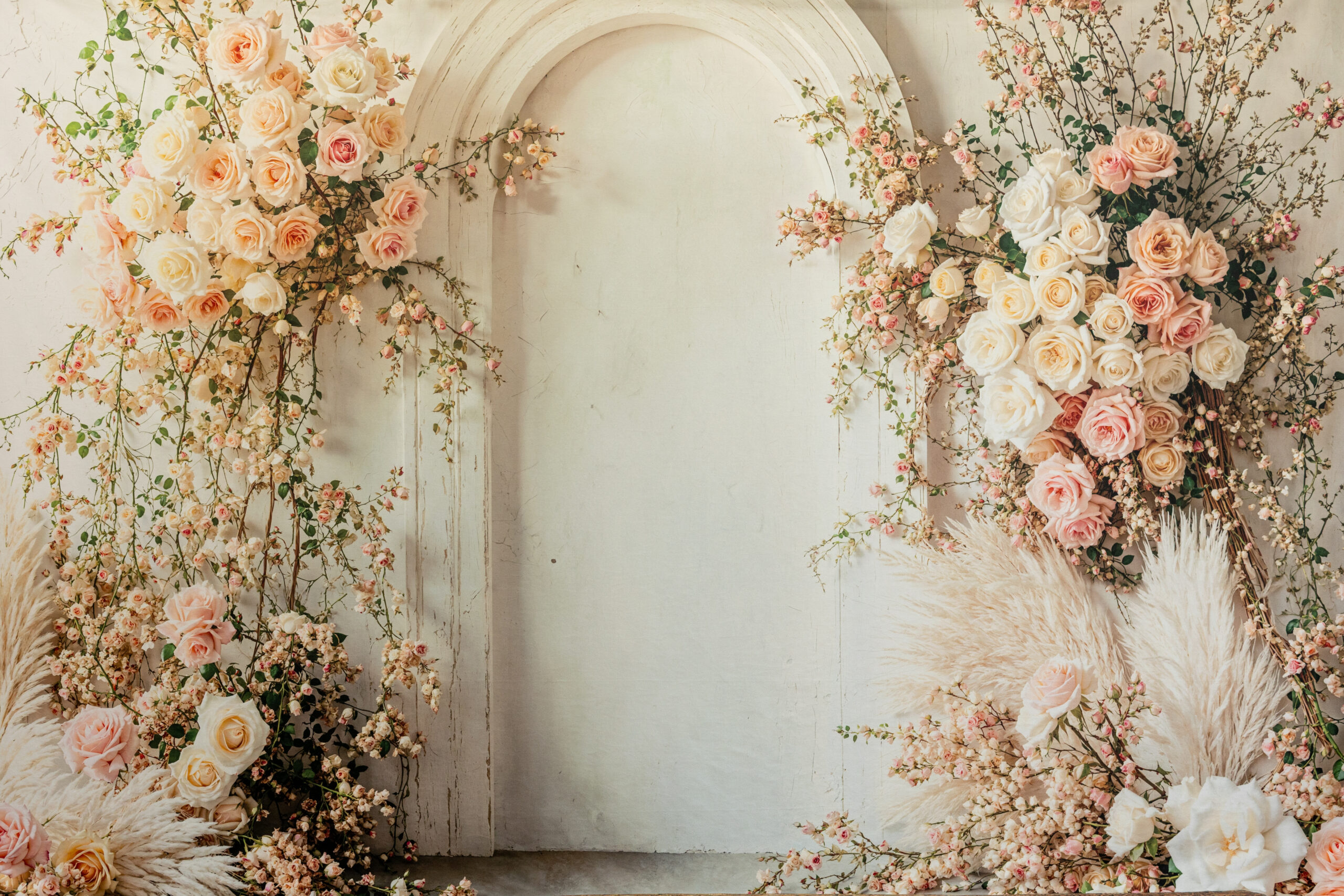 Vintage white arched doorway surrounded by lush floral arrangements featuring cream, blush, and soft pink roses, delicate trailing blossoms, and pampas grass, creating a romantic, garden-inspired backdrop with a shabby chic aesthetic.