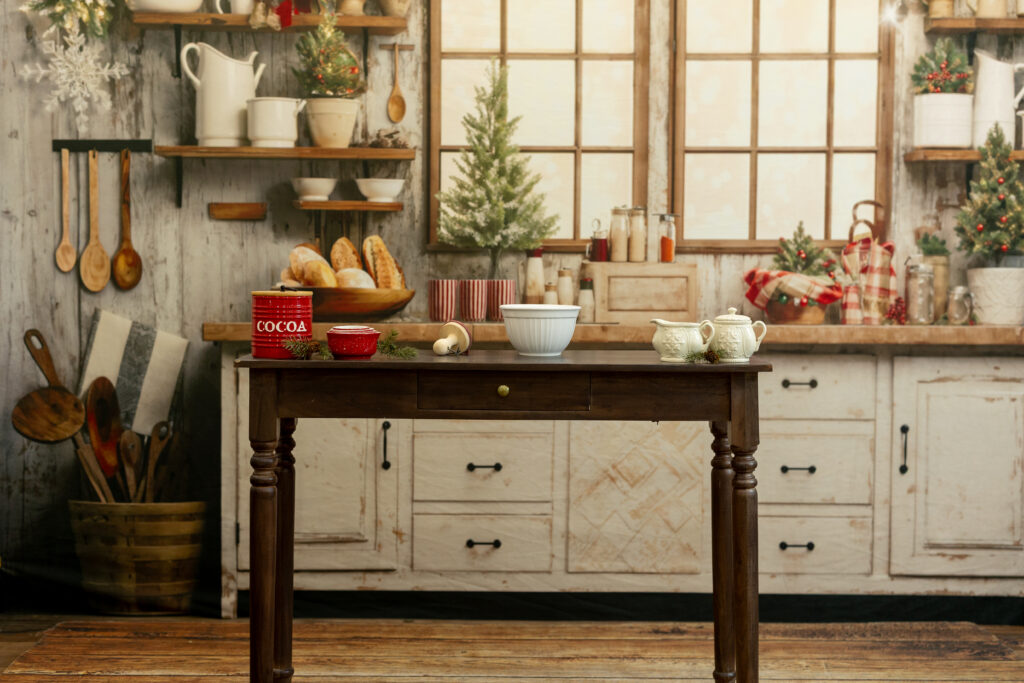 Rustic Christmas kitchen scene with a wooden table in the foreground holding holiday-themed items including a red cocoa tin, a small red cup, a white mixing bowl, and a cream-colored teapot. The background features vintage-style cabinets, shelves with kitchen tools and bread, small Christmas trees, and festive decorations.