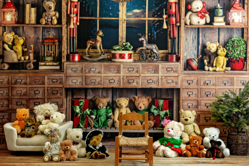 Whimsical Christmas scene featuring a wooden toy room backdrop filled with teddy bears, vintage drawers, rocking horses, nutcrackers, and festive decorations. A small wooden chair sits center stage in front of a white sofa and numerous stuffed animals arranged around a Christmas tree.