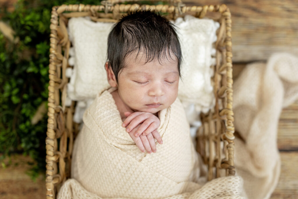 Close-up of a swaddled newborn in beige tones, showing tiny hands tucked under his chin.
