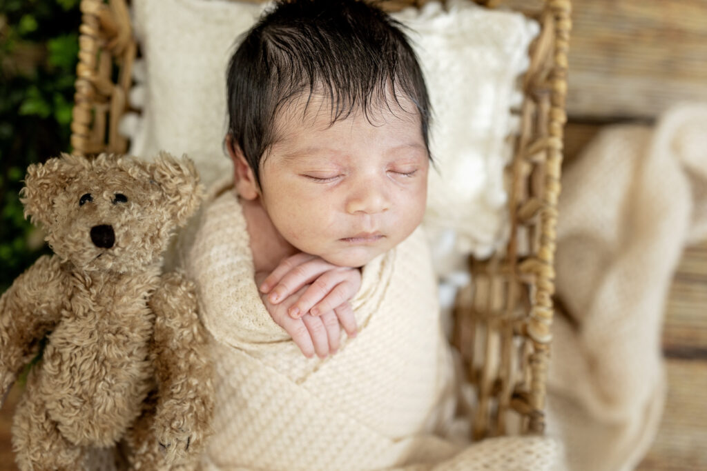 Newborn with arms gently folded near his face, wrapped in light neutral tones on wood backdrop