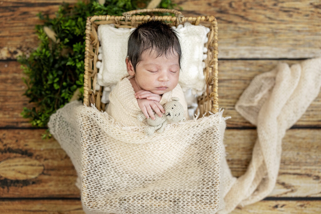 Baby boy in cream knit wrap, nestled in a cozy basket against rustic studio flooring.