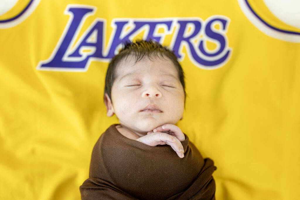 Newborn wearing a yellow Lakers jersey wrap, posed for a sports-themed photo.
